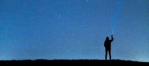 Figure of a man, standing against the backdrop of stars, shining a torch into the sky.