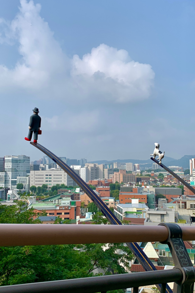 A pair of statues near Ehwa Mural Village in Seoul, South Korea. One of the statues is of a little man in a coat and bowler hat and the other is a white and black dog. They're extended over a walkway, overlooking South Korea's capital city: Seoul.