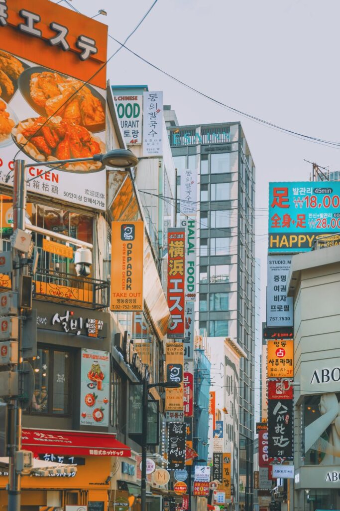Street photograph of Myeongdong in Seoul; known for being a shopping district in South Korea. There are billboards all along the street, selling products in Korean. The image is bright and slightly overwhelming - so many colours, tall buildings and advertisements in central seoul.